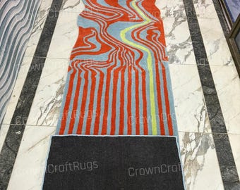 Op de afbeelding: Een abstract tapijt met een uniek ontwerp. Het tapijt heeft oranje, lichtblauwe en gele patronen. De onderkant van het tapijt is zwart. Het tapijt ligt op een marmeren vloer.