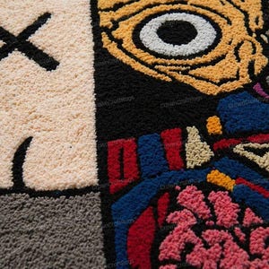 May include: Close-up of a colorful, textured rug with a cartoon-style design. The rug features a beige face with a black 'X' for eyes, a black mouth, and a large eye with yellow and white details. The design includes red, blue, and pink elements.