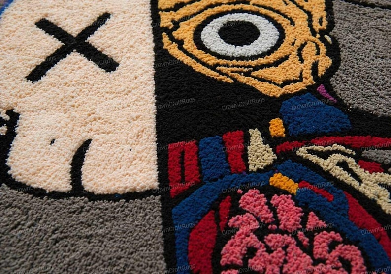May include: Close-up of a colorful, textured rug with a cartoon-style design. The rug features a beige face with a black 'X' for eyes, a black mouth, and a large eye with yellow and white details. The design includes red, blue, and pink elements.