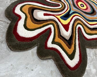 May include: An irregular, organic-shaped tufted rug featuring concentric, wavy bands of color including olive green, mustard yellow, white, deep red, black, light blue, and pink. The text "CrownCraftRugs" is visible in a repeating pattern across the rug.