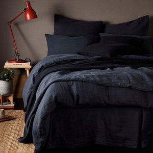 Puede incluir: Una cama con ropa de cama azul oscuro, que incluye una funda nórdica, almohadas y una falda de cama. Una manta azul oscuro está sobre la cama. Una lámpara de escritorio roja y una pequeña mesa auxiliar con una planta son visibles.