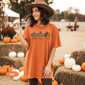 May include: A burnt orange short-sleeve t-shirt featuring a graphic of three pumpkins with leopard print patterns and a sunflower accent. The text "HAPPY FALL Y'ALL" is printed in dark brown letters below the pumpkins. The shirt has a crew neckline.