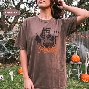 May include: An espresso brown short-sleeve t-shirt featuring a graphic of a pin-up style devil character with horns, bat wings, and a pitchfork. Below the character, the orange text reads Raisin' HELL. The shirt has a crew neckline and loose fit.