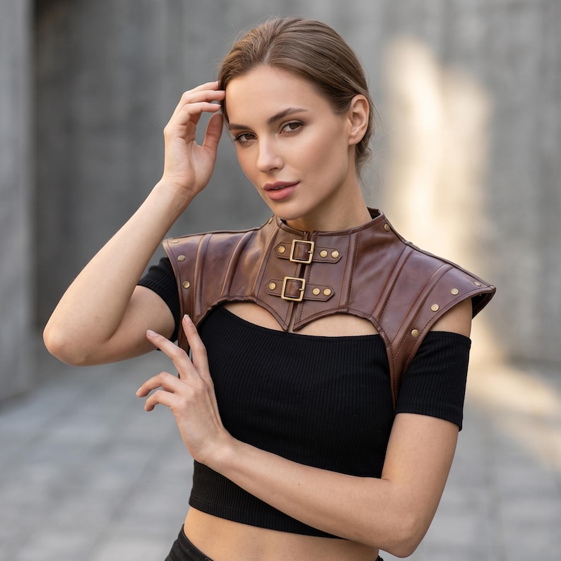May include: A brown leather-like shoulder armor piece featuring a high collar, two front buckle closures with brass-toned hardware, and decorative metal studs along the edges. The garment is worn over a black short-sleeved ribbed top.
