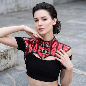 May include: A metallic red bolero-style shoulder piece featuring black trim, silver-toned metal studs, and two front buckle closures. The garment is constructed with vertical panels and a high collar, worn over a plain black short-sleeved crop top.