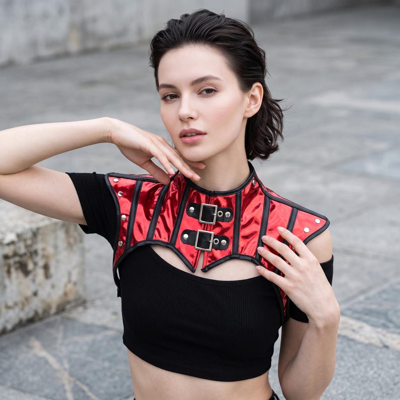 May include: A metallic red bolero-style shoulder piece featuring black trim, silver-toned metal studs, and two front buckle closures. The garment is constructed with vertical panels and a high collar, worn over a plain black short-sleeved crop top.