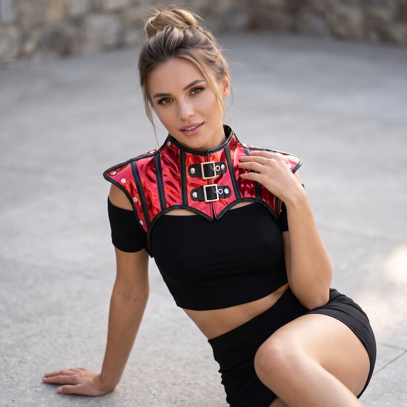 May include: A red metallic high-neck shoulder harness featuring black trim, silver-toned studs, and two front buckle closures. The harness is worn over a black short-sleeved crop top and a matching black skirt, creating a structured layered look.