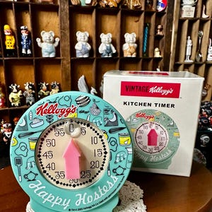 May include: A round, light blue Kellogg's Happy Hostess kitchen timer with a pink arrow indicator and a white dial featuring black numbers from 0 to 55. The timer is decorated with black line art of kitchen tools and food, and comes with a white box.