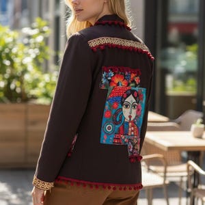 May include: A dark brown jacket with colorful embroidered patches and red pom-pom trim. The jacket features a detailed illustration on the back, with gold and red accents along the collar and hem. The jacket is worn with brown pants.