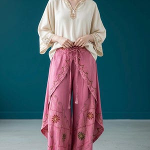 May include: A cream-colored V-neck blouse with gold embroidery on the neckline and sleeves, paired with pink wide-leg trousers featuring a wrap-style front, a drawstring waist, and intricate floral embroidery in gold and dark red thread.