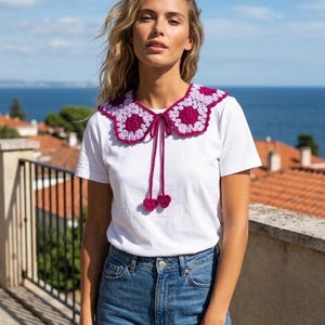 May include: A white t-shirt with a crochet collar. The collar is a combination of light purple and dark purple, with a tie and two heart-shaped accents. The model is wearing blue jeans.