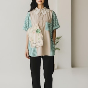 May include: A beige and green patchwork shirt with a lace collar and a floral embroidered pocket. The shirt has short sleeves and a button-down front. The person is wearing black pants and white sneakers.
