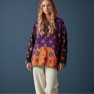 May include: A hooded pullover featuring a purple floral print on the upper body and sleeves, contrasted by an orange floral print kangaroo pocket. The garment includes a drawstring hood with pom-pom trim along the hem and sleeve cuffs.