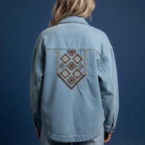 May include: A light blue denim jacket featuring a pointed collar, long sleeves, and a button-up front. The back panel displays a decorative embroidery pattern consisting of multiple diamond shapes in shades of brown, tan, and blue thread.