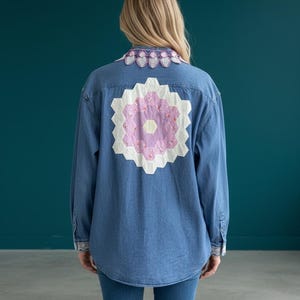 May include: A blue denim button-down shirt featuring a large, central hexagonal quilt patch on the back in shades of white, light purple, and floral pink. The collar and cuffs are accented with a decorative, scalloped lavender crochet trim.