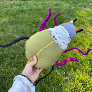 May include: A plush insect toy featuring a large olive green fabric body with a gold rickrack trim, a grey ribbed head, and a white eyelet lace collar. The creature has multiple long, thin, flexible legs in purple and black fabric with orange accents.