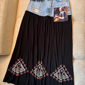 May include: A long skirt featuring a light blue acid-wash denim waistband with a small black tulle ruffle and a rectangular patch reading WEAR A-GAIN. The skirt includes a print of a painting and a black lower section with embroidered geometric patterns.
