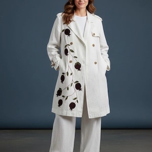 May include: A white double-breasted trench coat featuring a long, winding vine embroidery with dark brown pomegranate fruit and green leaves on the right side, paired with matching white wide-leg trousers and white sneakers.