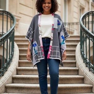 May include: A gray patchwork poncho with white grid pattern and floral accents. The poncho has a white trim and is worn over a white top and blue jeans. The woman is smiling and standing on a stone staircase.