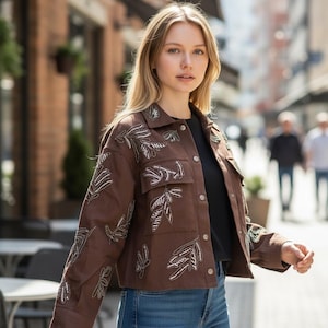 May include: A brown jacket with leaf-like embellishments. The jacket has two chest pockets and silver button closures. The leaf designs are made of white and silver beads. The jacket is worn over a black top and blue jeans.