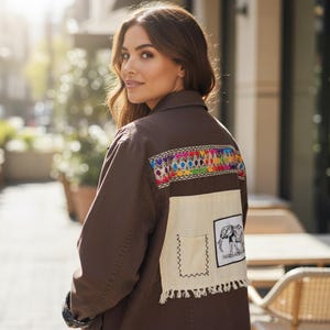 May include: Brown jacket with colorful embroidered detail across the upper back. The jacket features a cream-colored patch with an elephant graphic and fringe. The jacket is worn outdoors.
