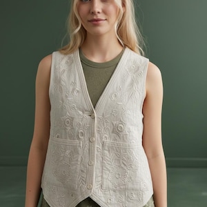 May include: A sleeveless beige waistcoat featuring intricate tonal floral embroidery throughout the front panels. The garment includes a V-neckline, a four-button front closure, and two square patch pockets on the lower front, all crafted in a light fabric.