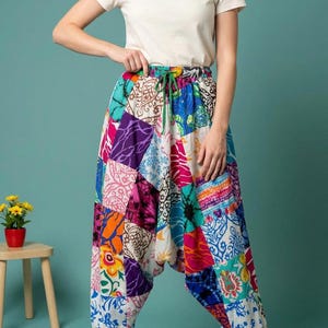 May include: A pair of loose-fitting harem pants featuring a vibrant patchwork design with various floral, tie-dye, and geometric patterns in shades of blue, pink, orange, purple, and white, finished with an elastic waistband and a green drawstring.