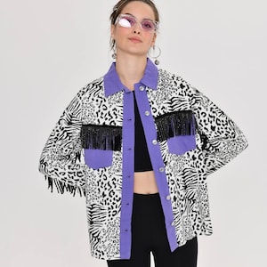 May include: A patterned jacket with a mix of zebra and leopard prints in black and white. The jacket features a purple collar and pocket accents, with black fringe detailing. It has a button-up front.