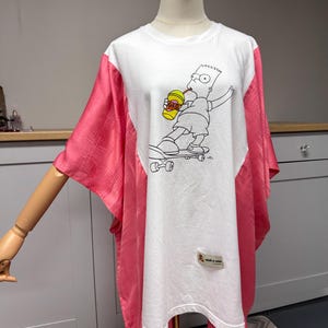 May include: White t-shirt with a graphic of Bart Simpson skateboarding and drinking a beverage. The shirt has pink sleeves and side panels. A small tag with text is visible on the bottom right.