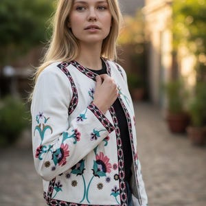 May include: White embroidered jacket with floral designs in pink, teal, and purple. The jacket features a black and pink patterned trim along the edges and sleeves. The jacket is paired with blue jeans.