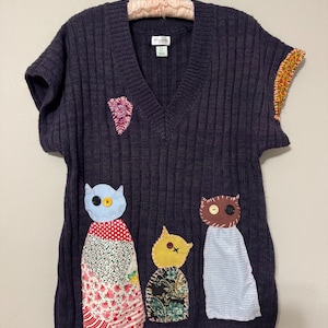 May include: Dark navy blue V-neck sweater with short sleeves, adorned with whimsical owl-like appliqués. The sweater features a ribbed texture and is embellished with colorful fabric patches, including floral and patterned designs. The sleeves have a decorative trim.