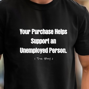 May include: A black short-sleeved t-shirt featuring white text that reads: Your Purchase Helps Support an Unemployed Person. Below this, in a smaller, handwritten-style font, the text reads: ( True Story ). The shirt has a crew neckline.