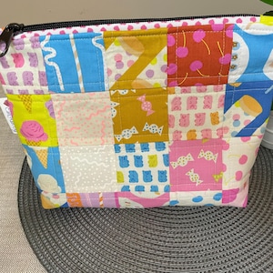 Puede incluir: Un colorido neceser de patchwork con cremallera negra. La bolsa presenta una variedad de cuadrados estampados con imágenes de conos de helado, ositos de goma y caramelos en tonos rosa, azul, amarillo y naranja.