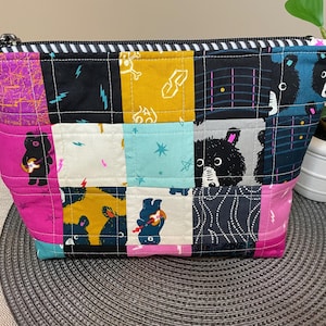 Puede incluir: Una bolsa con cremallera de patchwork con una variedad de cuadrados de tela coloridos. Los cuadrados presentan ilustraciones de osos, rayos y otros diseños. La bolsa tiene una cremallera a rayas blancas y negras.