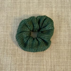 Może przedstawiać: Ciemnozielona gumka do włosów typu scrunchie o dzianinowej fakturze z widocznym pionowym prążkowaniem. Okrągły dodatek ma marszczony, elastyczny design, który tworzy miękki, objętościowy wygląd wokół centralnego otworu.