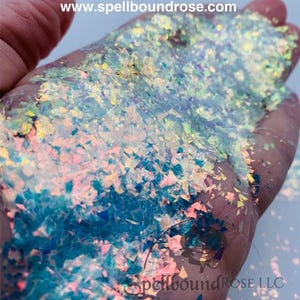 May include: A hand cradles iridescent opal glitter shards. The glitter, in shades of blue, pink, and yellow, has a reflective sheen. The image includes text: "Plié - Iridescent/Opal Glitter Shards" and "www.spellboundrose.com".