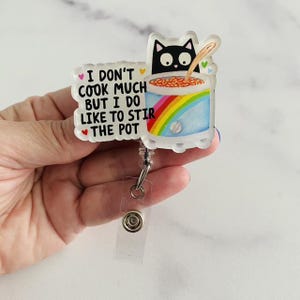May include: An acrylic badge reel featuring a black cat illustration behind a pot with a rainbow design. The text reads, I DON'T COOK MUCH BUT I DO LIKE TO STIR THE POT. The design includes small colorful hearts and a wooden spoon in the pot.