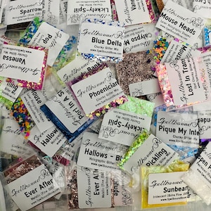 May include: A collection of small, clear plastic bags filled with colorful glitter. Each bag has a white label with product names like "Blue Delta" and "Phoenician" and the "Spellbound Rose" logo. The glitter colors vary.