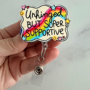 May include: A badge reel featuring a cream-colored, cloud-shaped acrylic topper with a rainbow graphic and the black text "Unhinged BUT SUPER SUPPORTIVE". The design includes small colorful stars, hearts, and yellow smiley faces around the edges.