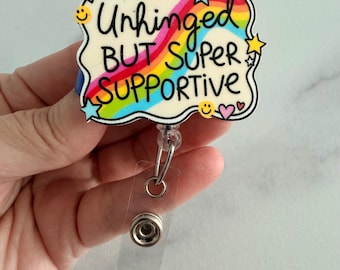 May include: A badge reel featuring a cream-colored, cloud-shaped acrylic topper with a rainbow graphic and the black text "Unhinged BUT SUPER SUPPORTIVE". The design includes small colorful stars, hearts, and yellow smiley faces around the edges.