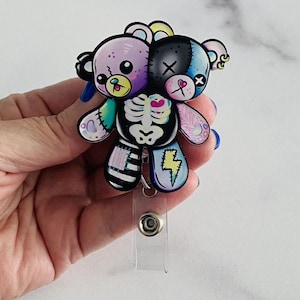 May include: A badge reel featuring a split-faced teddy bear design. One side is purple, the other is blue and black. The bear has a skeleton torso, stitched patches, and a lightning bolt. The reel has a clear plastic strap and a metal clip.