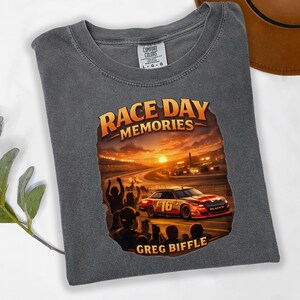 May include: Gray t-shirt with a graphic featuring a race car on a track at sunset. The graphic reads "Race Day Memories" and "Greg Biffle". The shirt is a Comfort Colors brand.