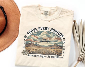 Könnte beinhalten: Ein cremefarbenes T-Shirt mit einem Grafikdruck eines Flugzeugs über einer Wüstenlandschaft und einer Startbahn. Der Text lautet ABOVE EVERY HORIZON und Adventure Begins At Takeoff, ergänzt durch einen Kompass, USA-Stempel und US-Flagge.