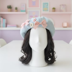 Peut inclure: Un béret bleu clair orné de nœuds roses et d'une fleur blanche est présenté sur une tête de mannequin avec des cheveux noirs ondulés. Le béret est décoré de plusieurs petits nœuds roses et d'une fleur blanche.
