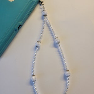 May include: A white beaded phone strap with six pearl-like balls. The strap is attached to a teal phone case.