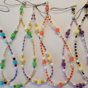 May include: Six colorful phone charms with smiley face beads and crystal beads. The charms are made with different colored beads, including purple, green, orange, and black.