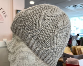 May include: A gray knitted hat with a cable knit pattern. The hat is displayed on a mannequin head.
