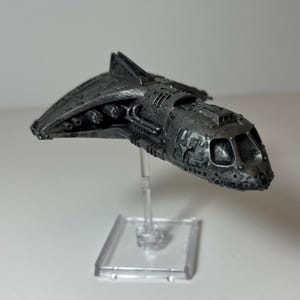 Peut inclure: Maquette de vaisseau spatial gris métallique détaillée, avec des détails de surface complexes. Le vaisseau est monté sur un support transparent, mettant en valeur son design. Le modèle présente un cockpit et divers éléments mécaniques.