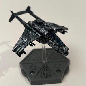 Può includere: Un modello in miniatura di aereo grigio scuro con ali, motori e cabina di pilotaggio dettagliati. Il modello è montato su una base esagonale grigia con segni incisi. L'aereo ha un design futuristico.