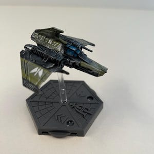 Op de afbeelding: Een gedetailleerd miniatuur ruimteschipmodel in groen- en blauwtinten. Het model is gemonteerd op een grijze zeshoekige basis met een transparante standaard. Het ontwerp suggereert een verzamelobject of een spelonderdeel.